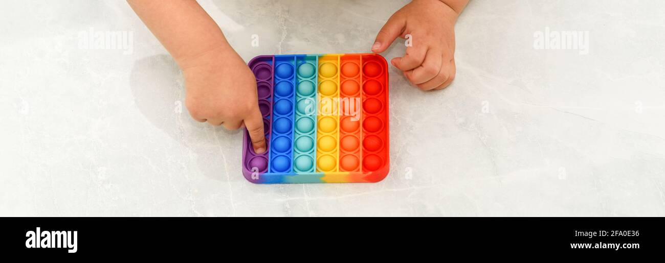 Anonymous child with colorful poppit game. Close up bunner shot of kid hands playing with colorful pop It fidget toy. Stock Photo