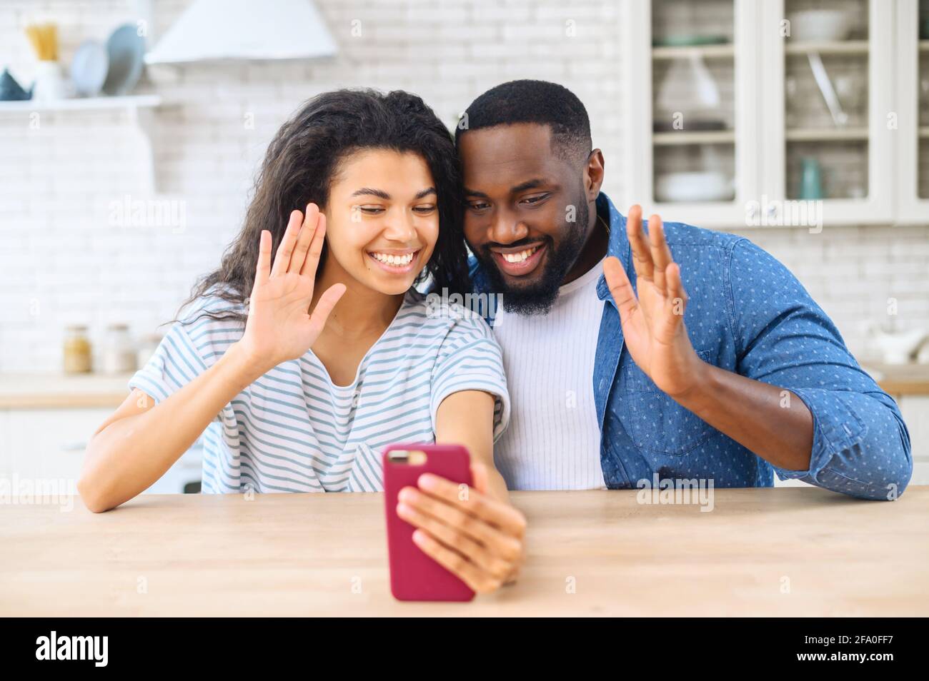 Front view of happy cheerful young multiracial couple having online webcam  chat with via video call on mobile phone, sitting together in the kitchen  in the cozy apartment. Concept of household life