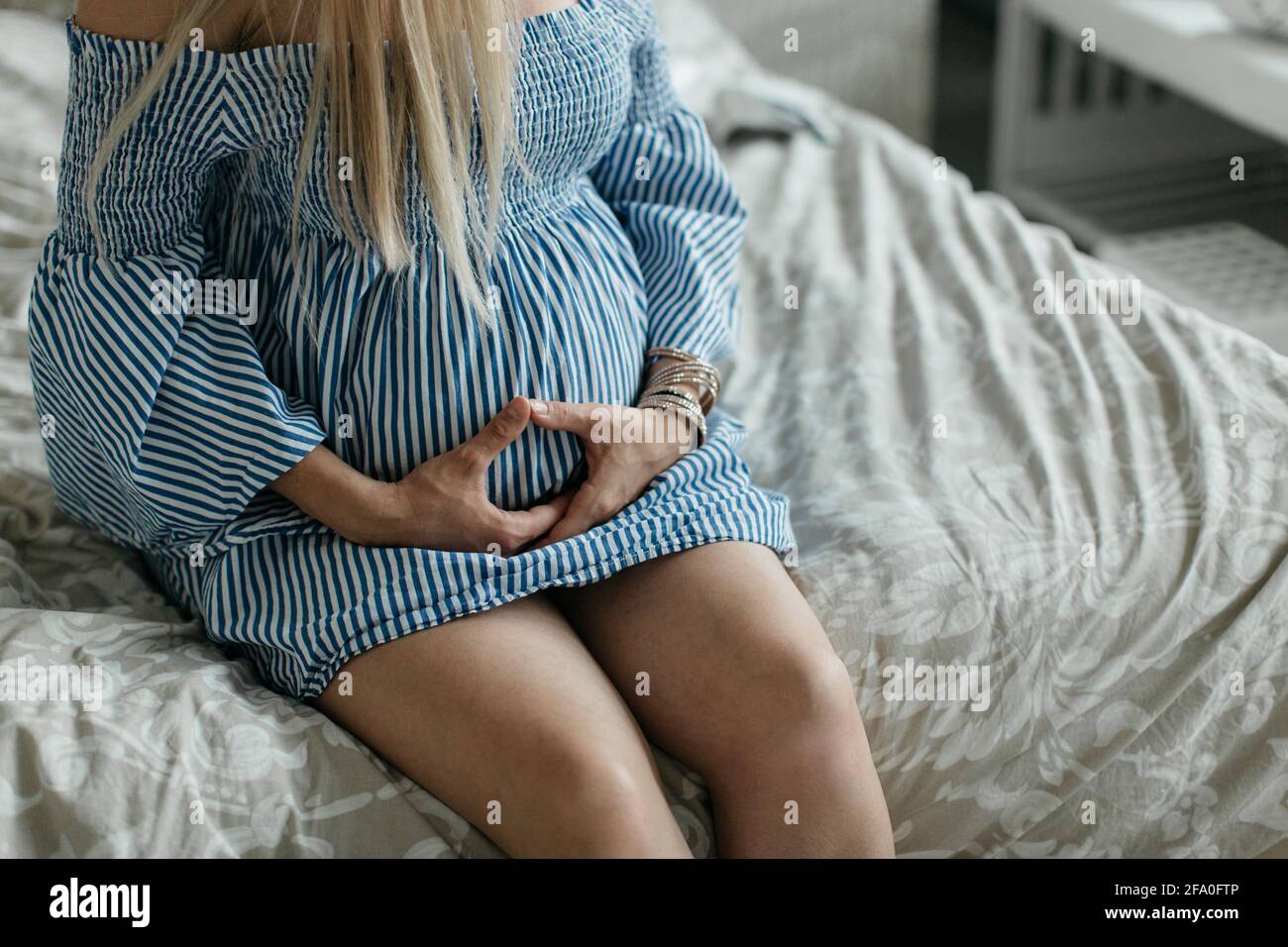 A cropped image of a pregnant woman sitting on a bed in a bedroom. A portrait of an expectant mother holding her belly. Stock Photo