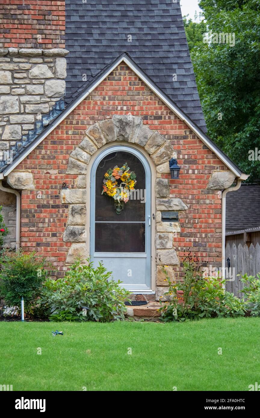 Cute brick and rock cottage with gabled entrance and arched door with bright green lawn and fall wreath on door. Stock Photo