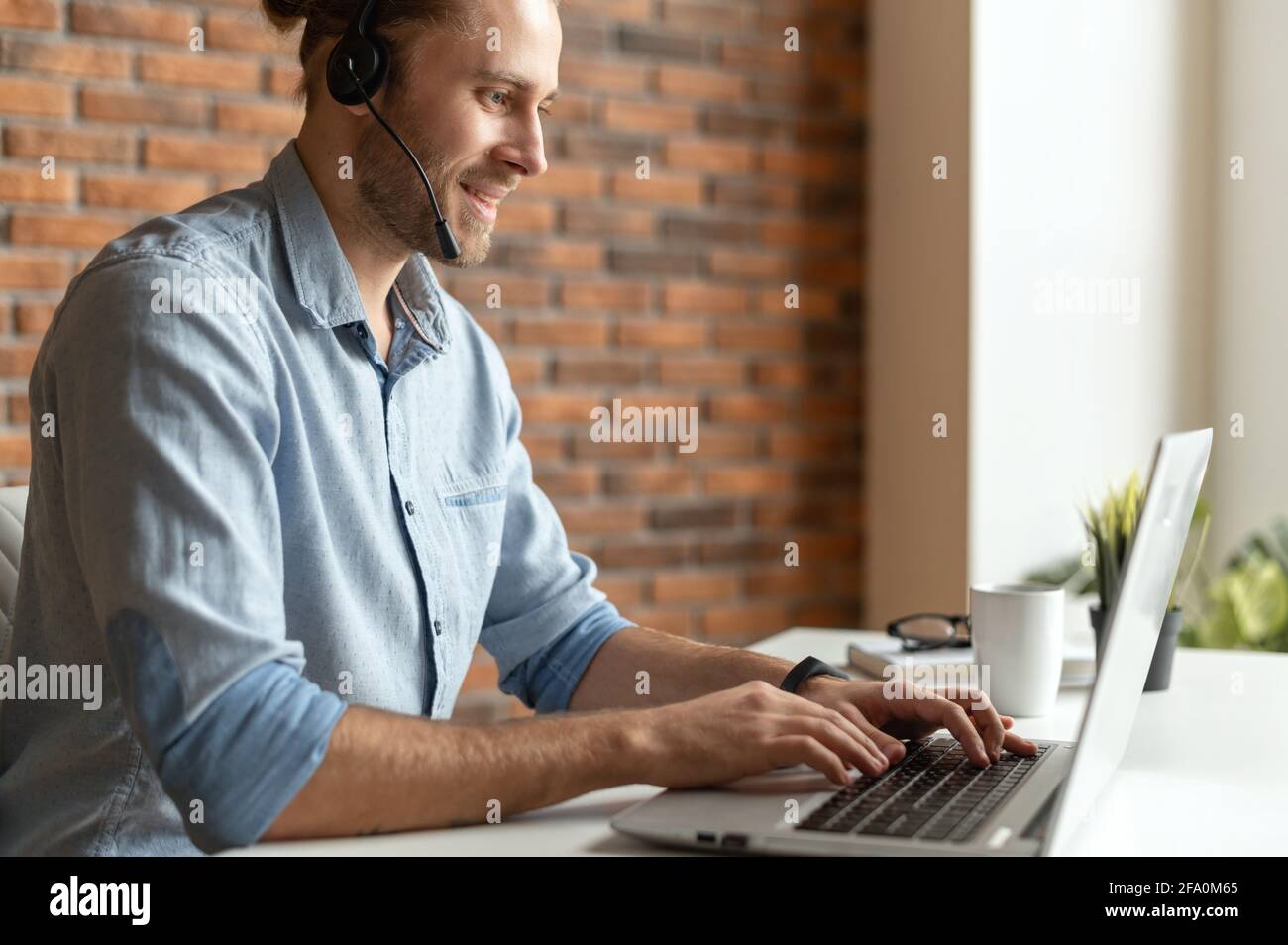 A confident male office worker sitting at the desk with a laptop, using headset communicating with customers, a helpful support officer speaking online and makes marks on computer app, side view Stock Photo