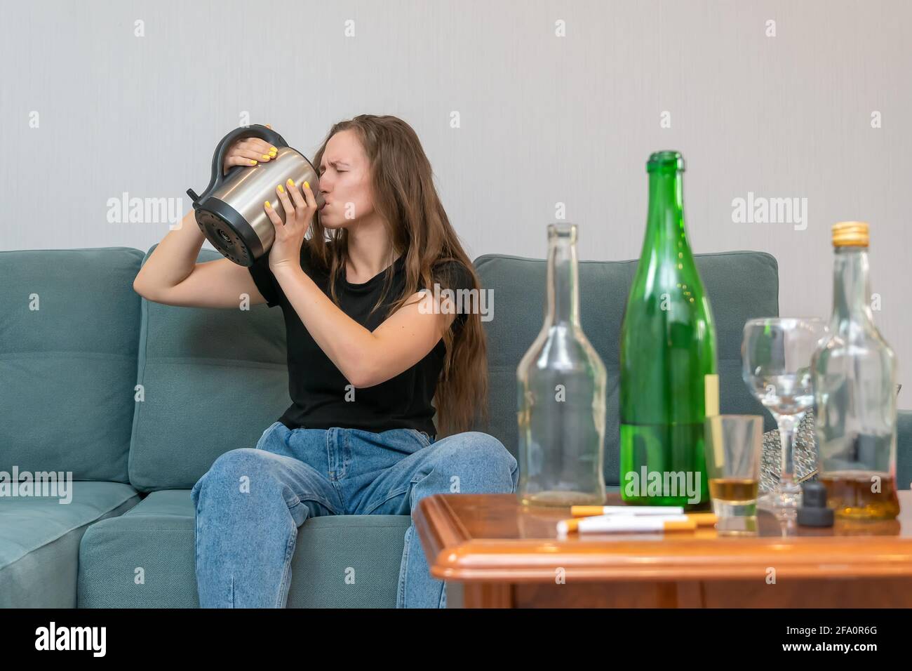 Young dark-haired woman sits on a sofa and drinks water from a teapot after a stormy party, empty bottles stand next to her Stock Photo