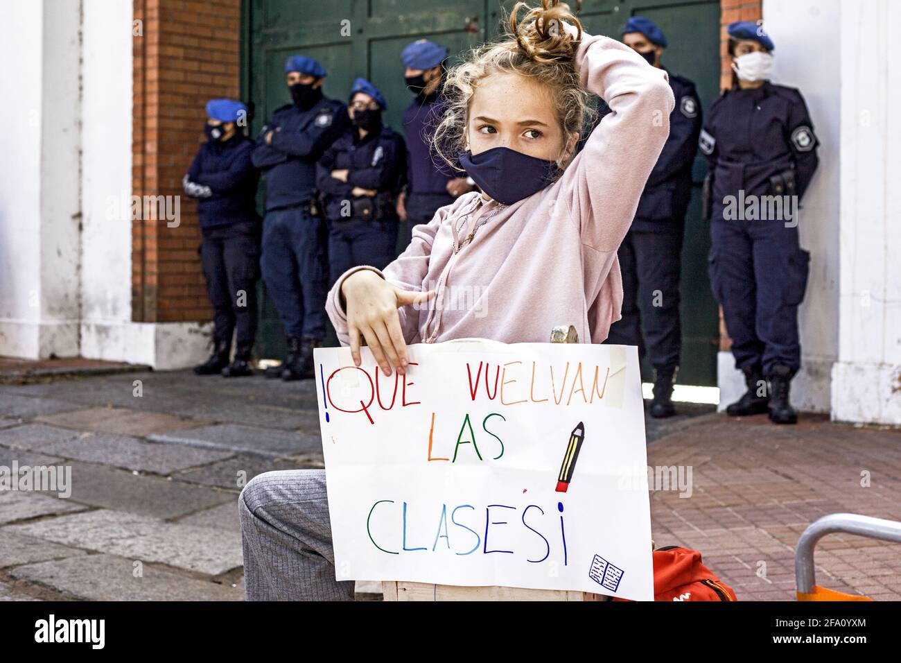 Buenos Aires, Federal Capital, Argentina. 21st Apr, 2021. A seated girl with police officers behind her stretches after finishing her homework outdoors in the vicinity of the Presidential residence. In front of her is a banner that reads: ''Que vuelvan las clases'' (Let the classes return). With Argentine schools closed, a group of parents organized a class in front of the Presidential Residence in protest against the suspension of classroom attendance for 15 days, a measure that President Alberto Fernandez ordered last Wednesday through a Decree of Necessity and Urgency to contain the resurg Stock Photo