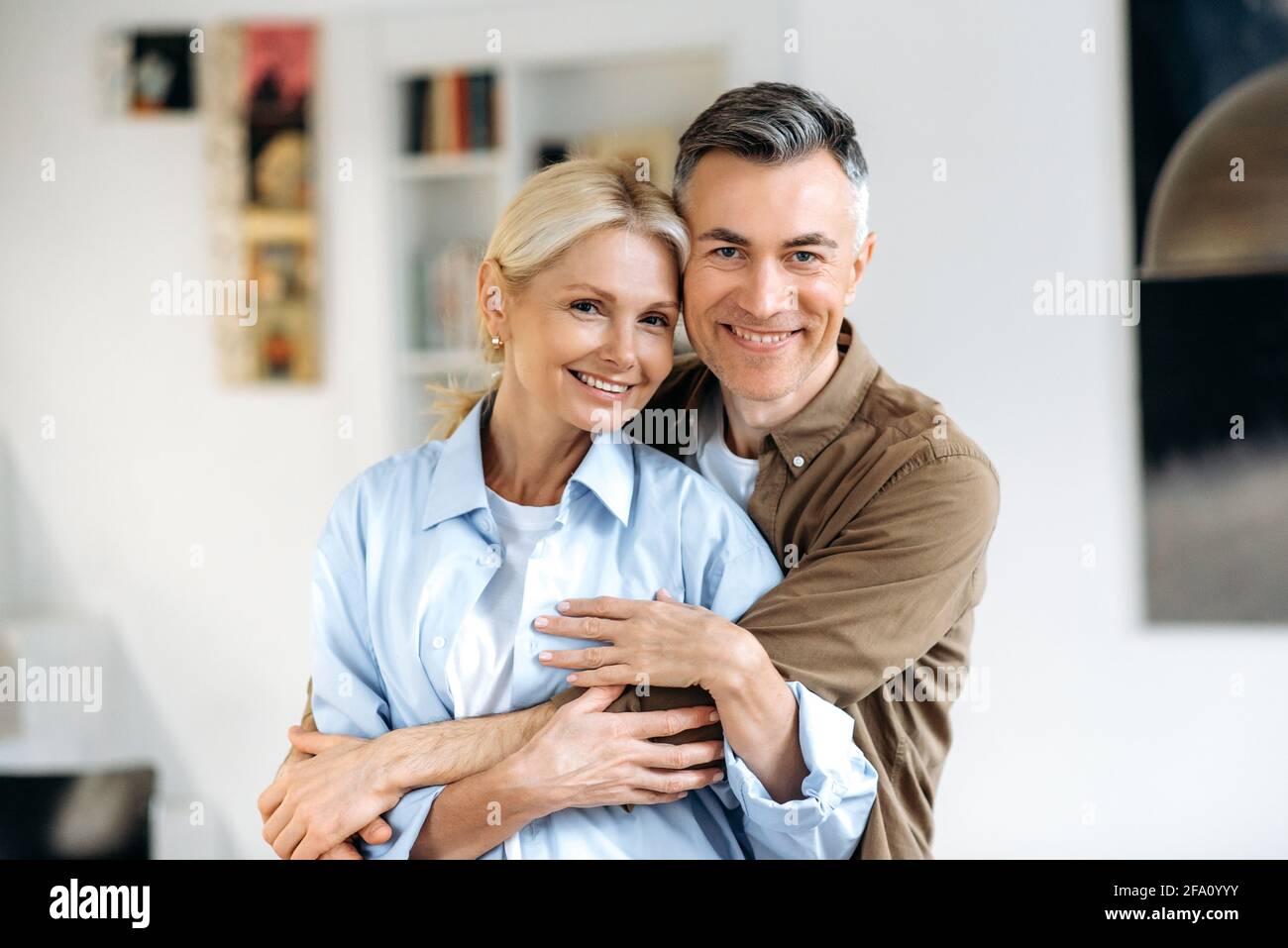 Family portrait. Close-up portrait of joyful happy mature caucasian married couple, stylishly dressed, husband and wife standing in living room, gently hugging each other, looking at camera, smiling Stock Photo