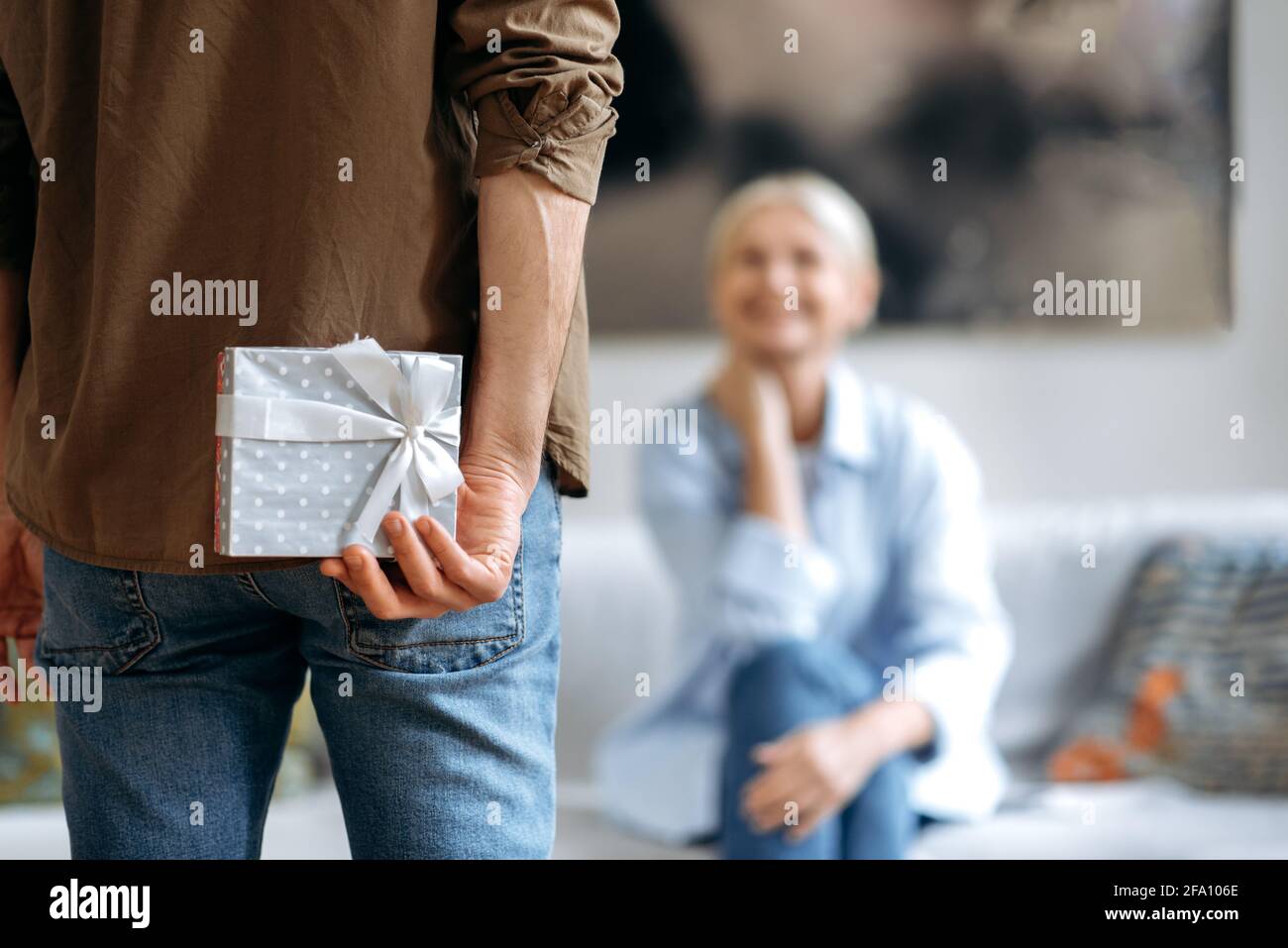 A present from husband on a special day. A loving husband in casual stylish clothes, holds a beautiful small box behind his back, prepares to give a surprise his beloved wife, a small anniversary gift Stock Photo