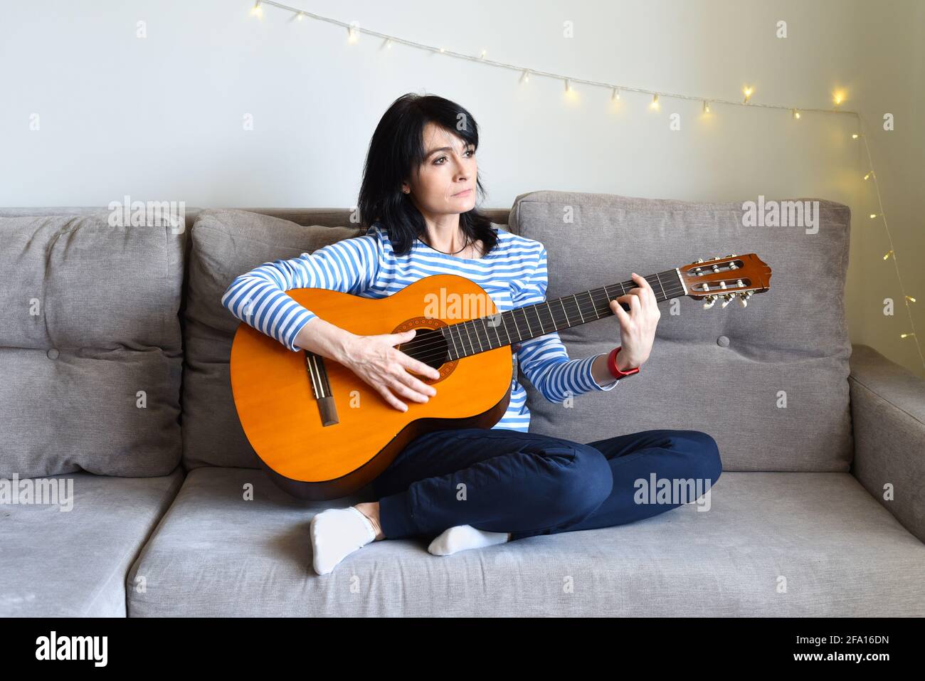45 y.o. Russian woman playing guitar, sitting on a sofa at home. Stock Photo