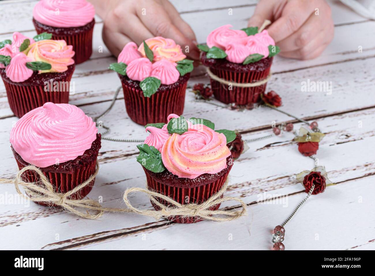Red velvet cupcakes decorated with sisal yarn bow, next to a female necklace. In the background, confectioner decorating a cupcake. Stock Photo