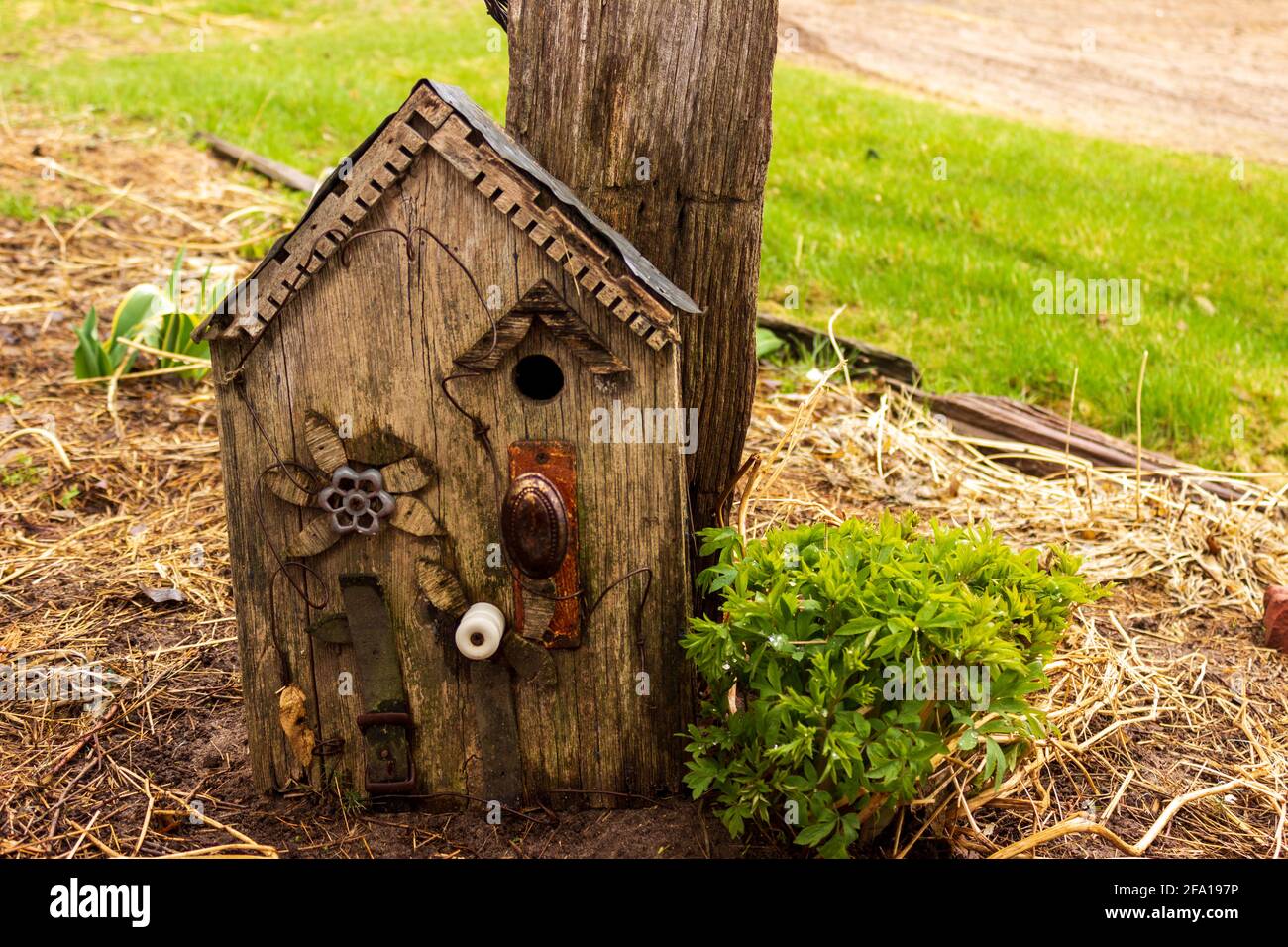 A rustic birdhouse made from a repurposed door handle, water faucet handle, belt, insulator, tin and old decorative wood leans against a post. Stock Photo