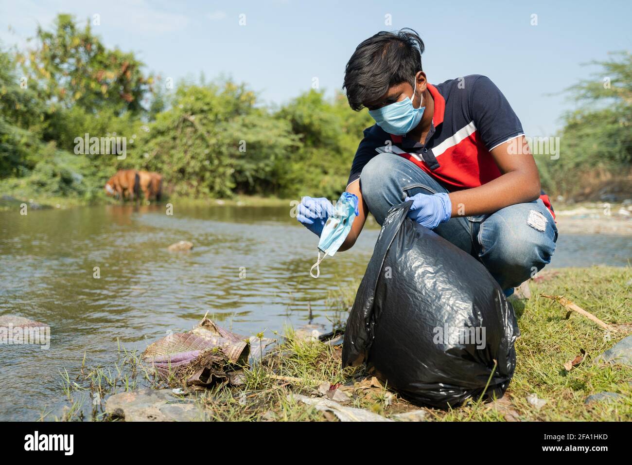 frontline worker or volunteer collecting trapped discarded face mask near lake or river water and placing inside garbage bag - concept of medical Stock Photo