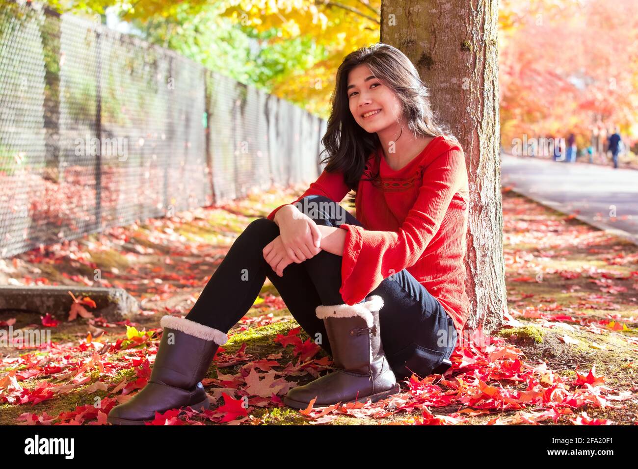 Smiling biracial teen girl in red shirt sitting under colorful autumn leaves of maple tree at park outdoors in fall season Stock Photo