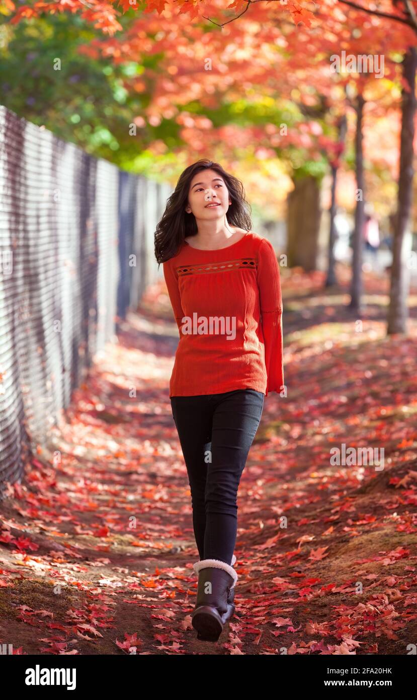 Smiling biracial teen girl in red shirt smiling, walking under red maple tree in park in autumn Stock Photo