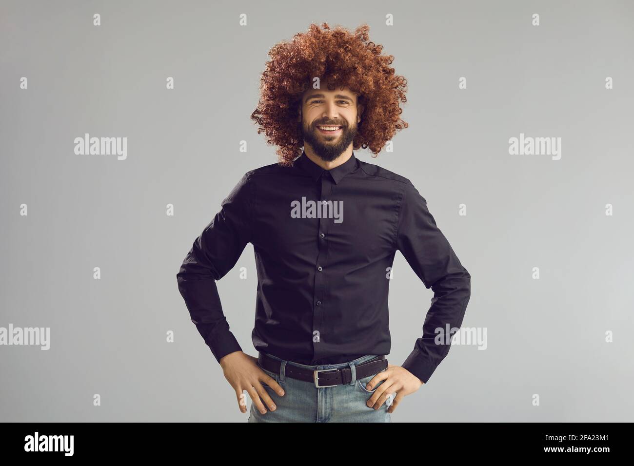 Funny happy man wearing curly brown wig standing hands oh hips on gray background Stock Photo