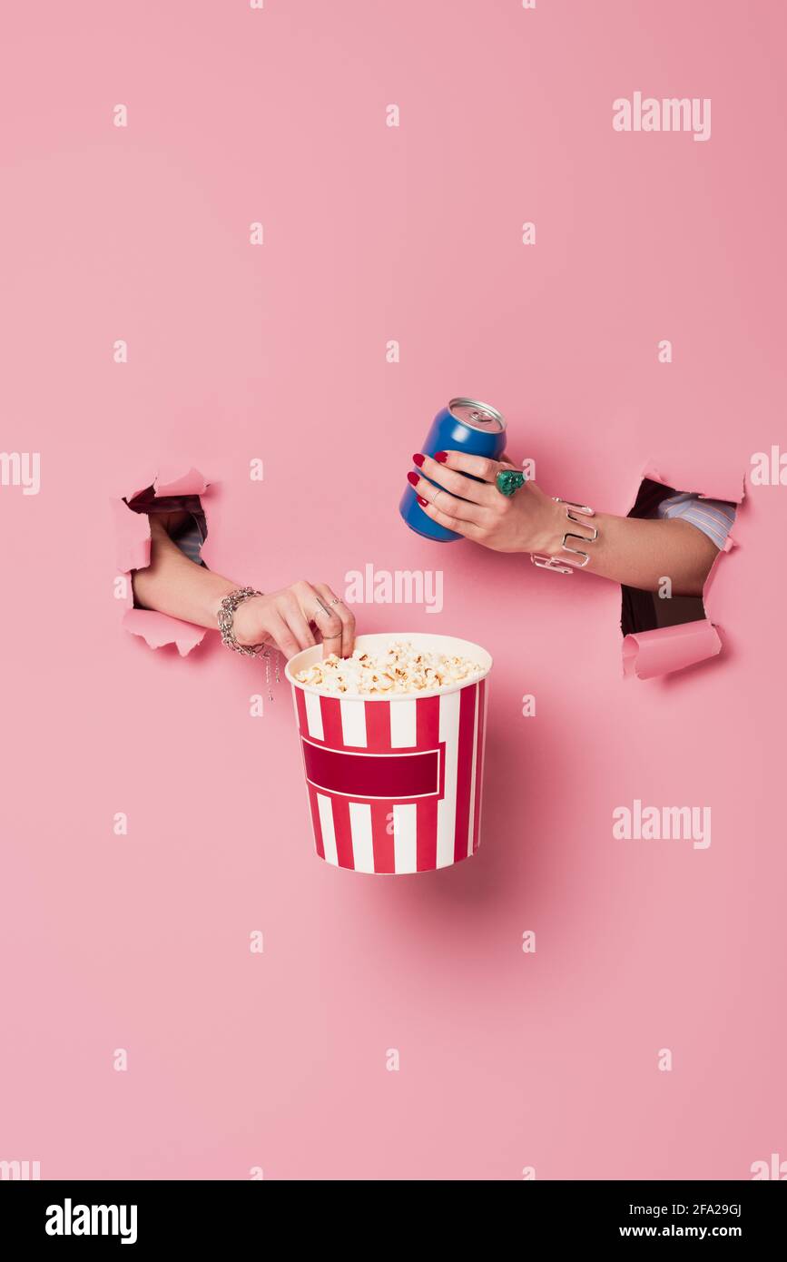 Cropped view of woman holding popcorn and drink in can near pink background with holes Stock Photo