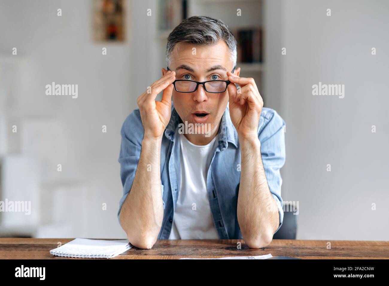 Surprised shocked handsome senior gray-haired caucasian man in casual  stylish clothes, sitting at table, lowering glasses with both hands, amazed  looking at computer webcam Stock Photo - Alamy