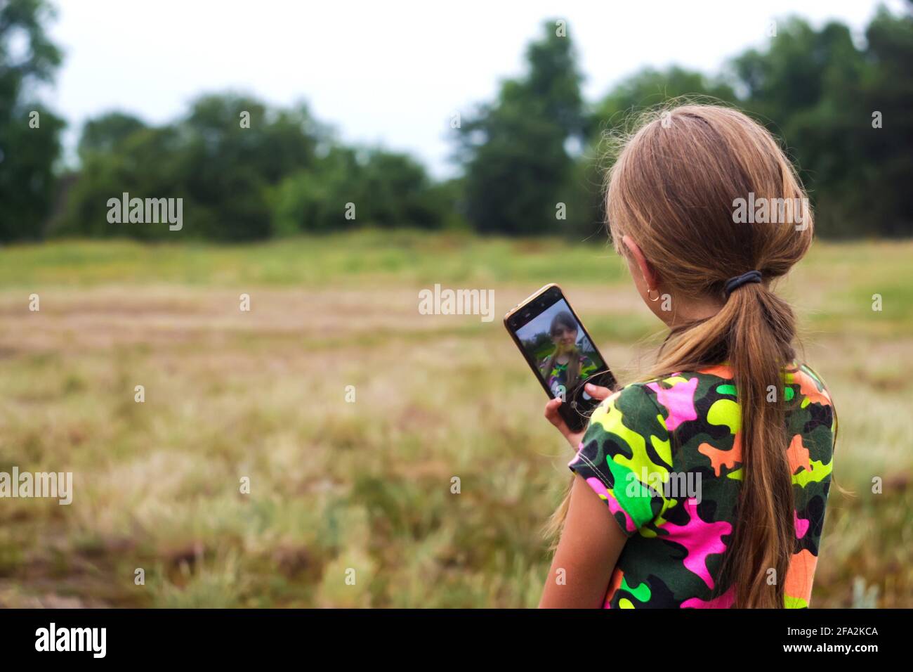 Defocus Selfie girl. Little girl takes a selfie by hand. Reflection on screen. Back view of preteen girl in summer standing on the meadow and taking a Stock Photo