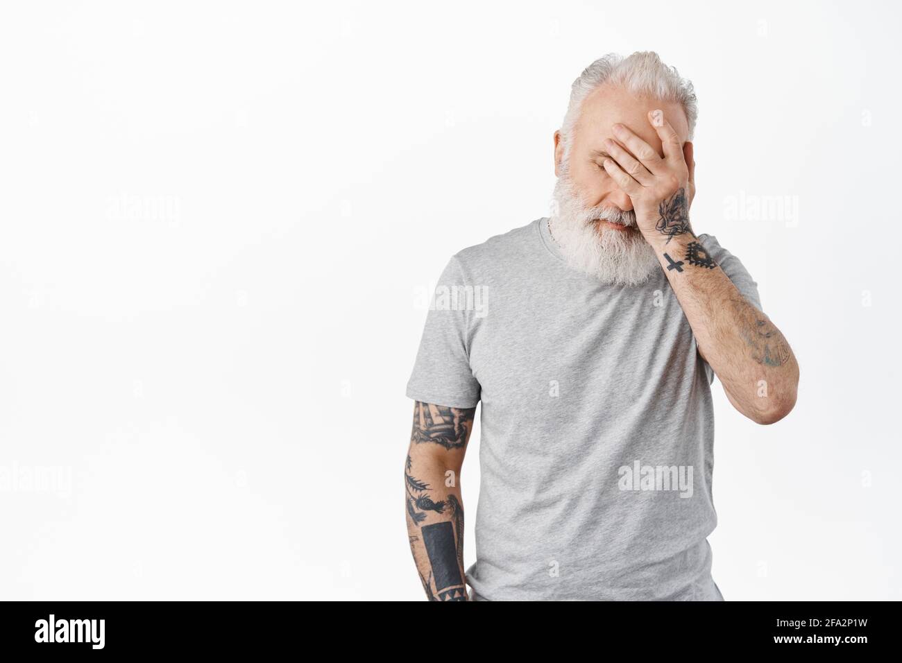 Oh gosh. Tired and annoyed senior man facepalm, hold hand on face distressed or exhausted, standing irritated in casual grey t-shirt against white Stock Photo