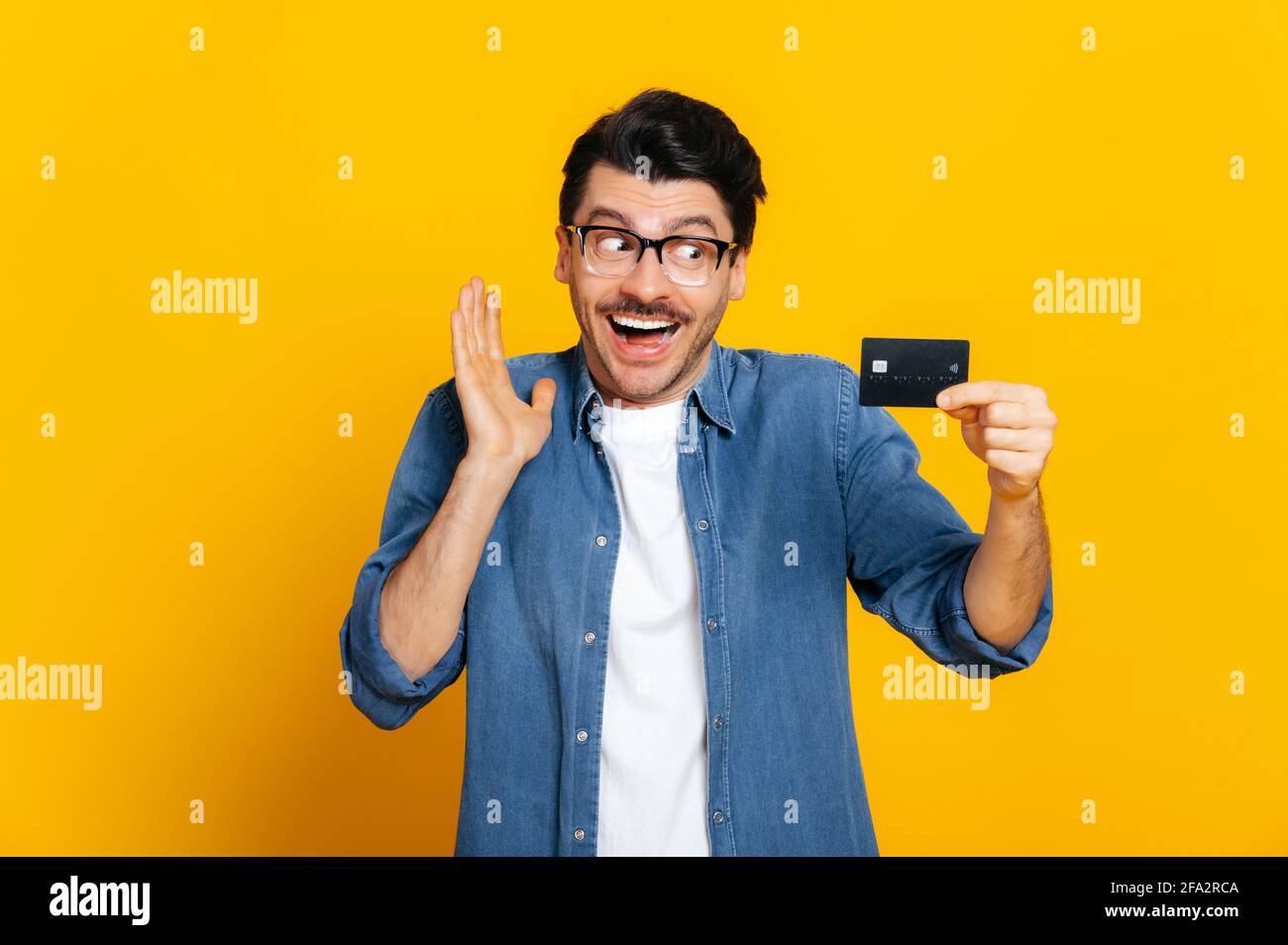 Happy excited caucasian unshaven handsome stylish guy with glasses, holding credit card in hand, looking at her in amazement, standing on an isolated orange background Stock Photo