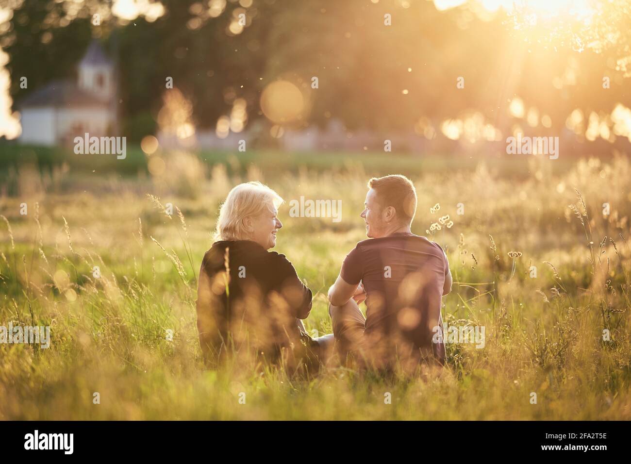 Mother sitting with son on meadow. Senior woman and young adult man talking against village at sunset. Stock Photo