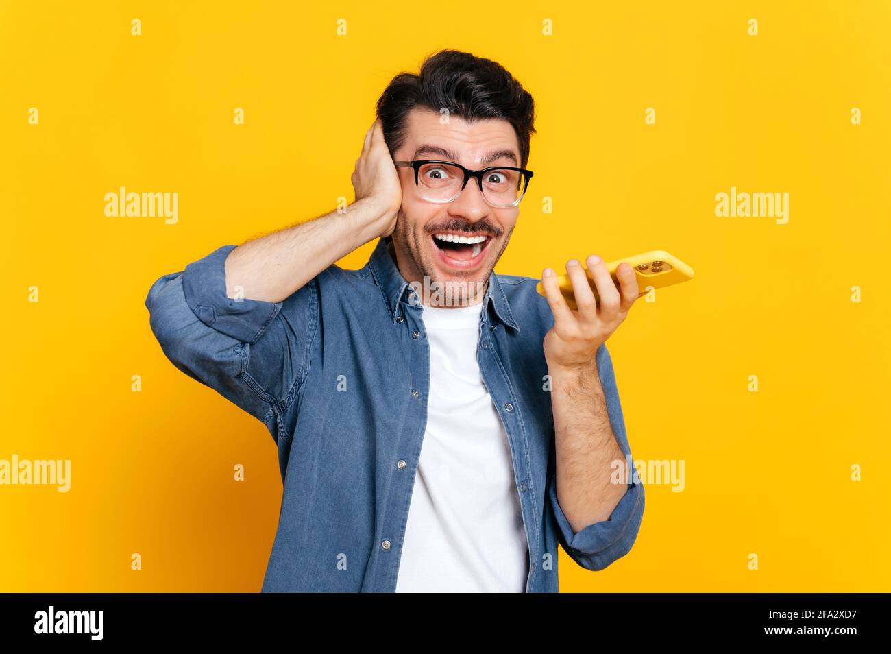 Emotional caucasian guy with glasses, hold smartphone in hand, screaming during phone call or recording audio message, stand on isolated orange background, holds his hand near head. Audio messaging Stock Photo