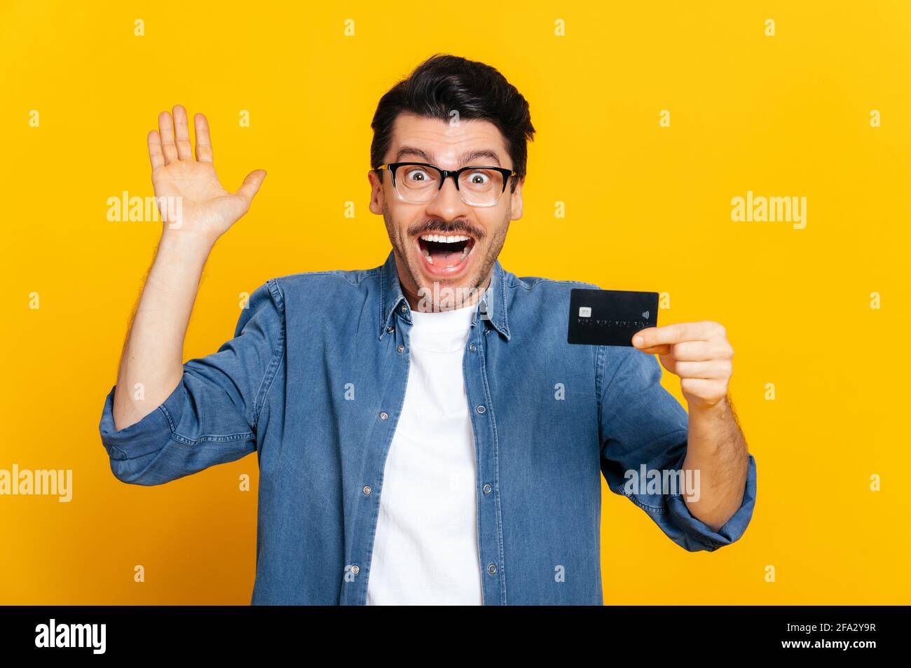 Young excited positive caucasian stylish man with glasses, holds credit card, rejoices in success, victory, profit, standing on isolated orange background happily raises his hand and smiling Stock Photo