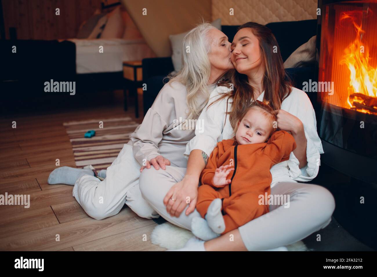 Grandmother, Mother and child kissing and playing on sofa near fireplace. Mom and baby. Parent with daughter and grandson little kid relaxing at home. Family having fun together. Mother's day Stock Photo