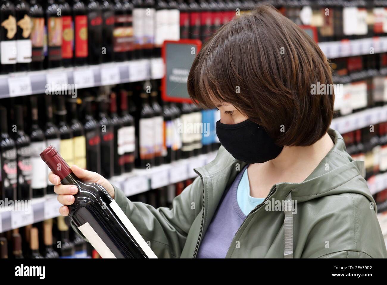 Woman in medical face mask reading the label on red wine bottle. Customer in liquor store, concept of choosing and buying alcohol Stock Photo