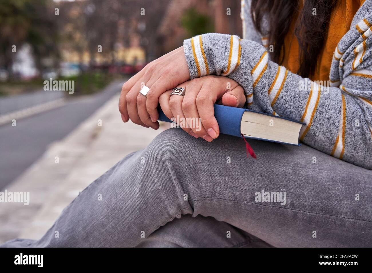 Hands crossed with rings in her fingers on closed book. Midsection detail of lady sitting in a park holding the book on her legs. Knowledge and read c Stock Photo