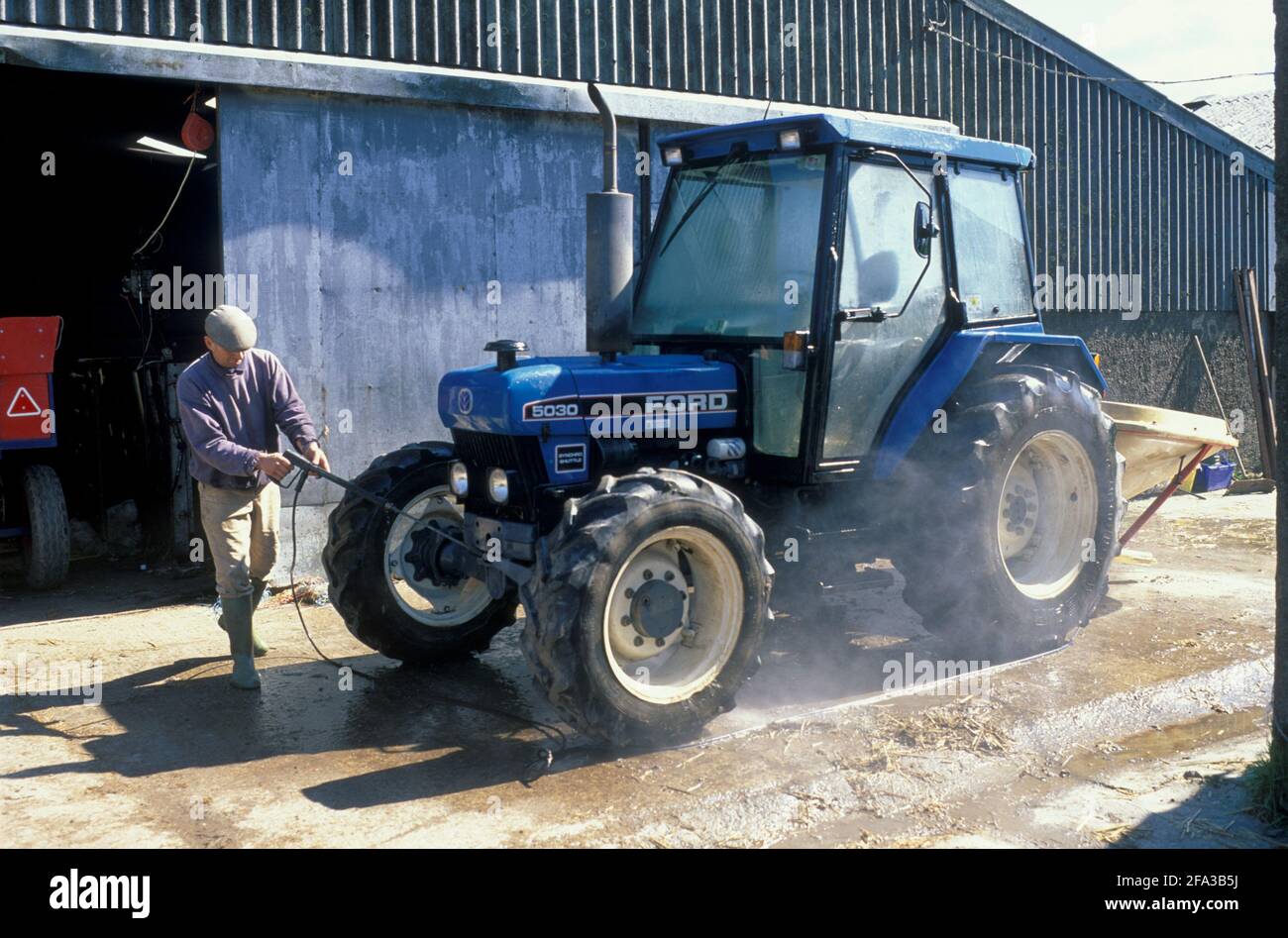 A Devon farmer washing his Ford 5030 tractor with a power-jet washer, on a hard concrete standing. Stock Photo