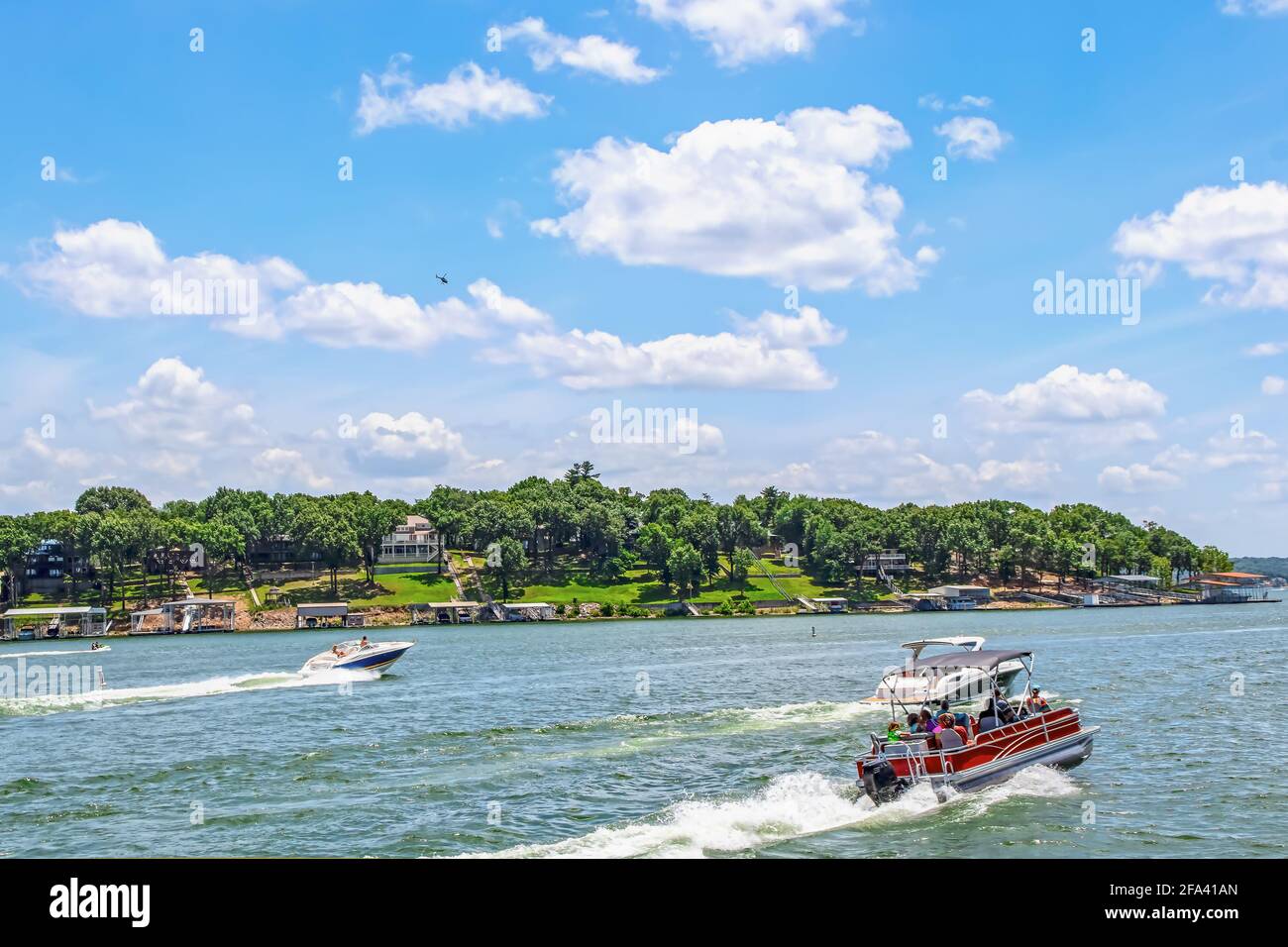 Pontoon boat full of people and two speedboats race down lake with luxury homes and docks on shore under bright blue sky with clouds and helicopter ab Stock Photo