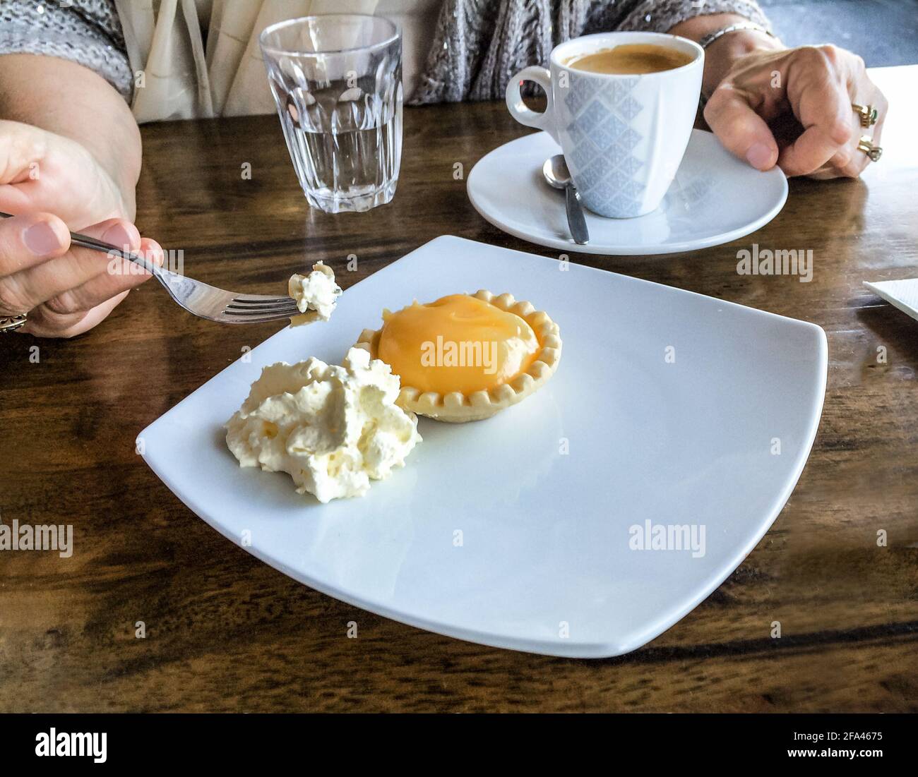 Woman's hand holding fork with bite of Lemon tart and clotted cream -coffee cup setting on table beside plate Stock Photo