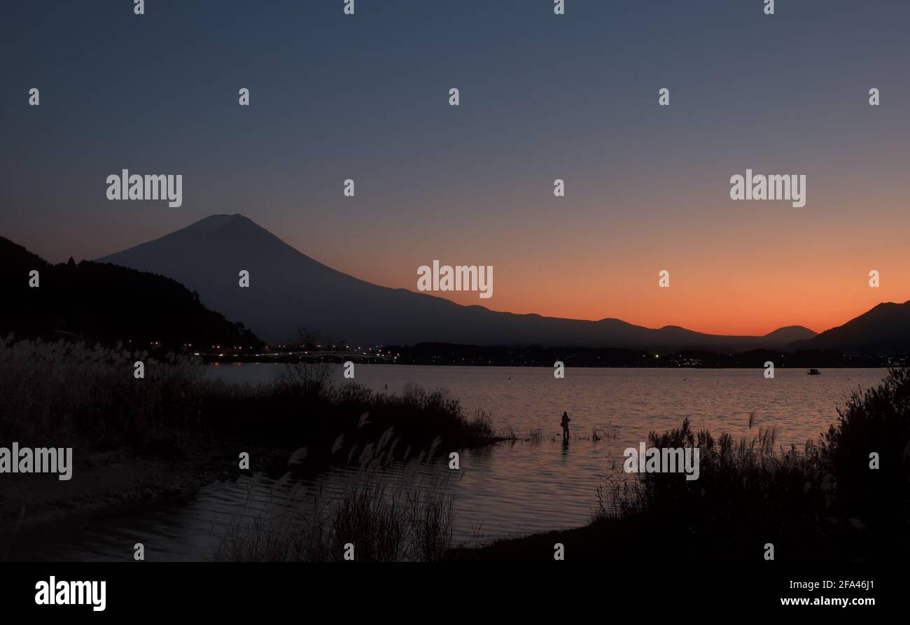 Early evening view of Mount Fuji and Lake Kawaguchi under a thin crescent moon, with a lone fisherman visible near the shore Stock Photo