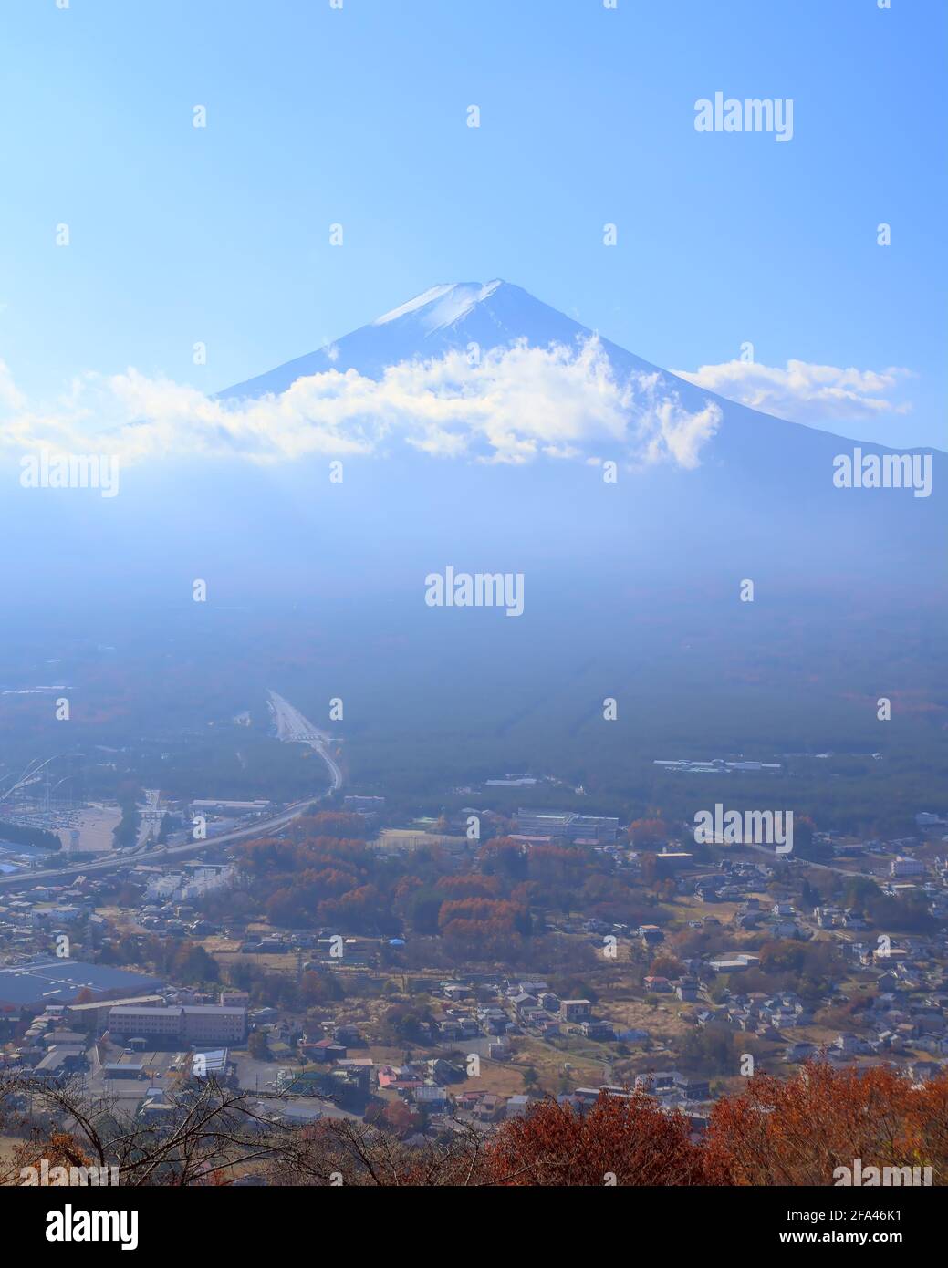 Daytime view of Mount Fuji from afar, partially covered by low clouds under a bright blue sky and with part of the city of Fujiyoshida visible Stock Photo