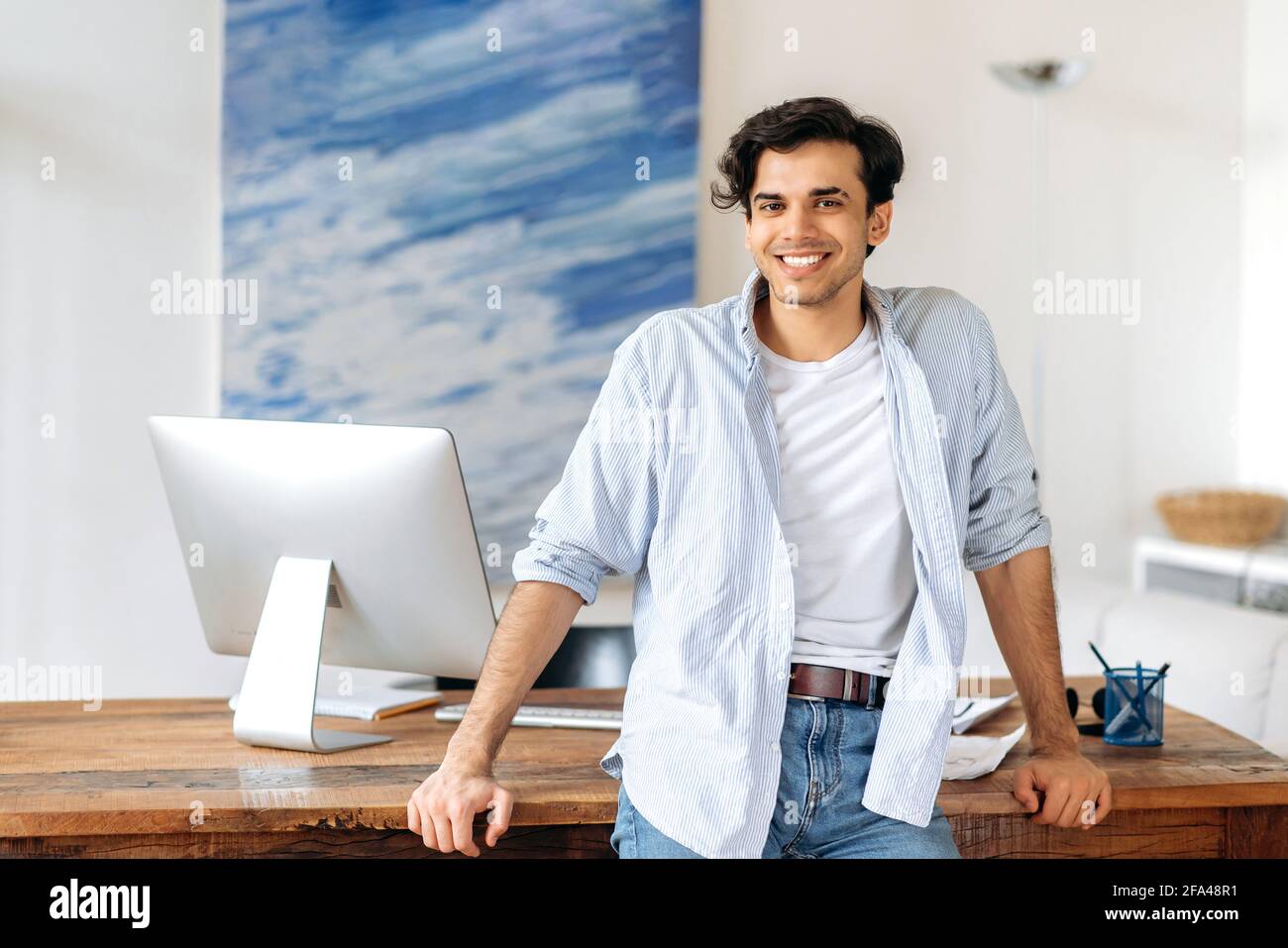 Portrait of confident successful young hispanic businessman, manager or freelancer, dressed in stylish clothes, standing near work desk, looking at camera, smiling friendly Stock Photo