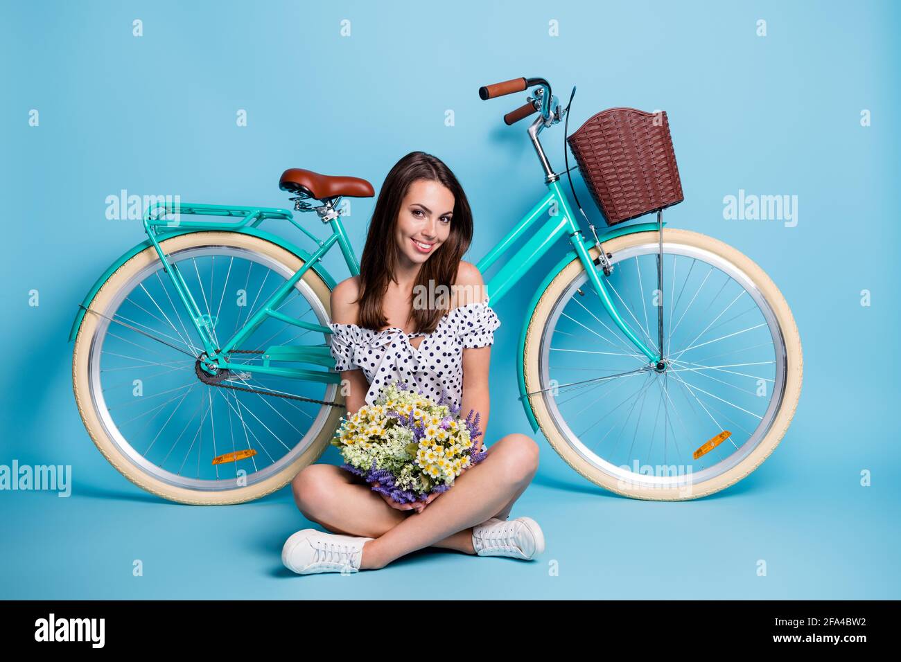 Full length photo of lady sit floor crossed legs hold bunch flower bike wear dotted dress footwear isolated blue color background Stock Photo