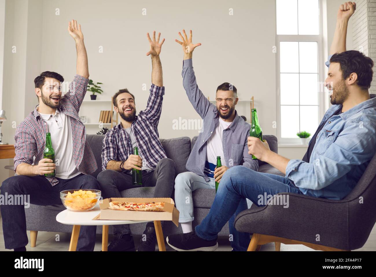 Smiling excited men group drink beer eat show hands up sit on home sofa Stock Photo
