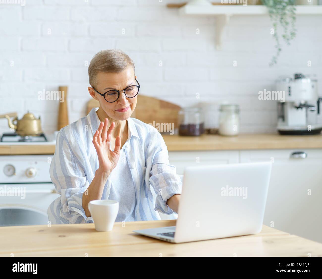 Modern smiling mature woman wearing glasses waving at webcam, studying  online on laptop while sitting in kitchen at home and drinking morning  coffee. Senior people and distance education concept Stock Photo -