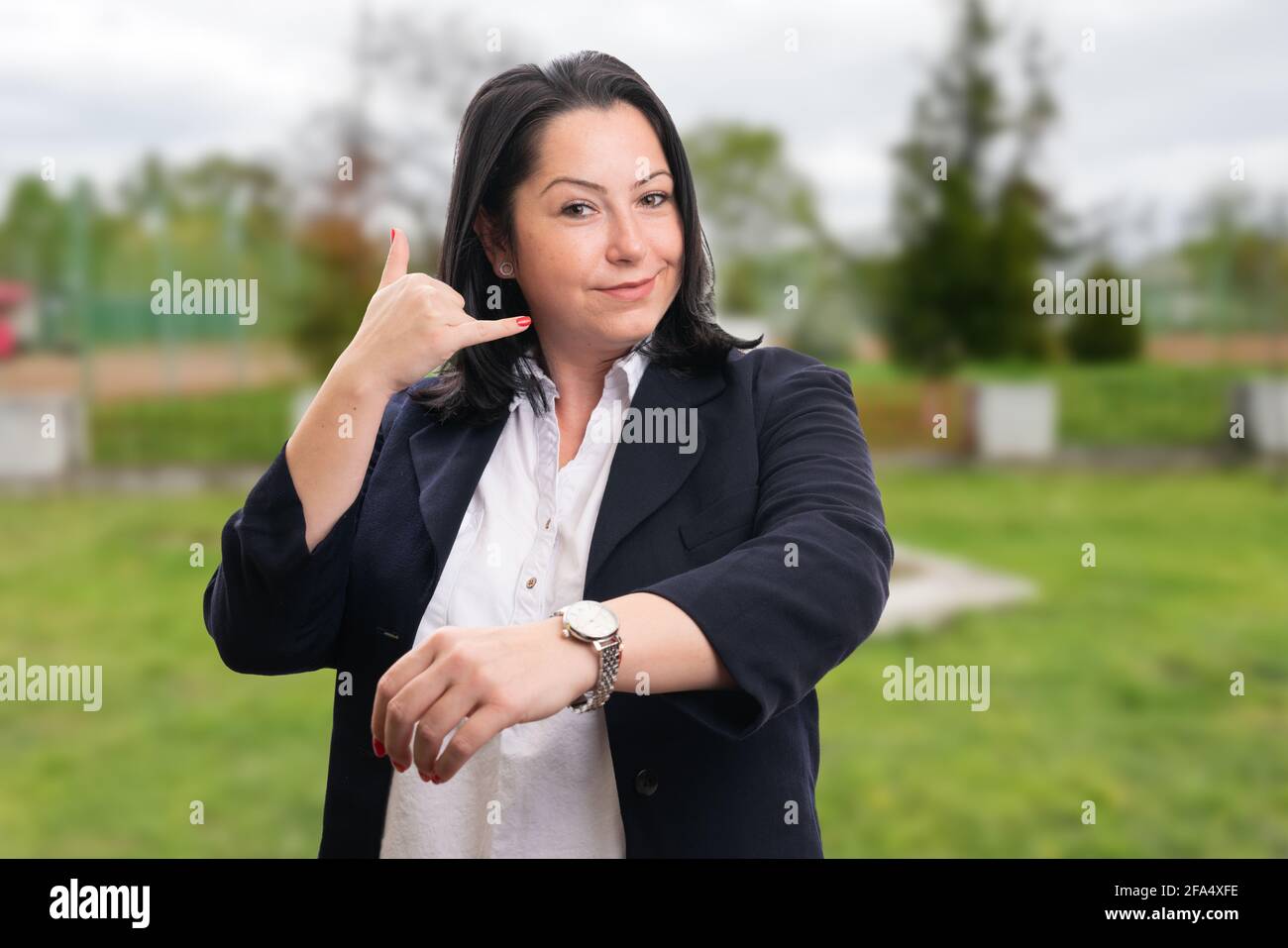 Corporate woman entrepreneur wearing suit smiling making calling gesture with fingers showing watch on wrist as telephone marketing concept in park na Stock Photo