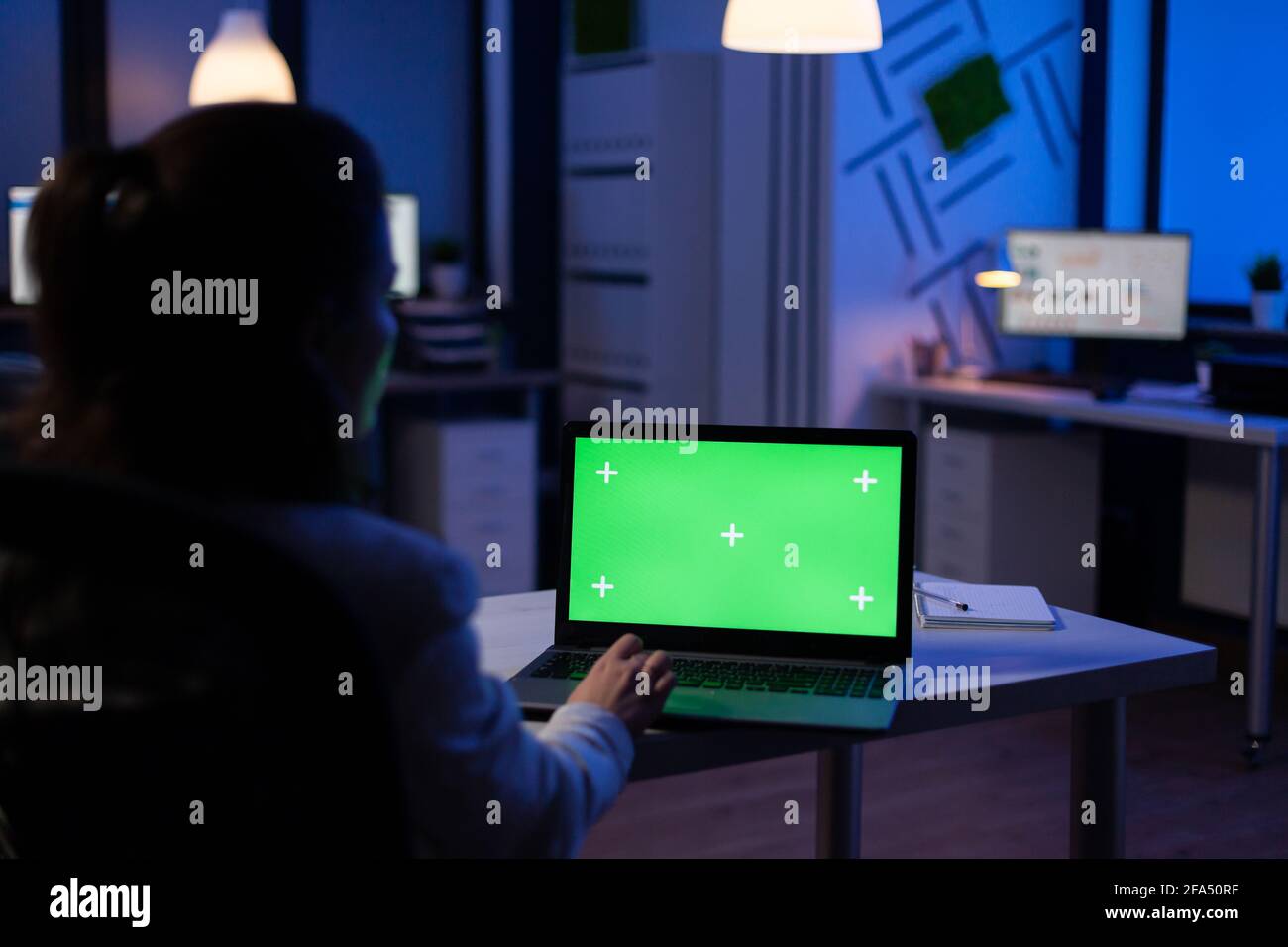 Back view of business woman looking at green screen laptop sitting at desk in start up company office . Exectutive manager watching desktop monitor display with green mockup, chroma key Stock Photo