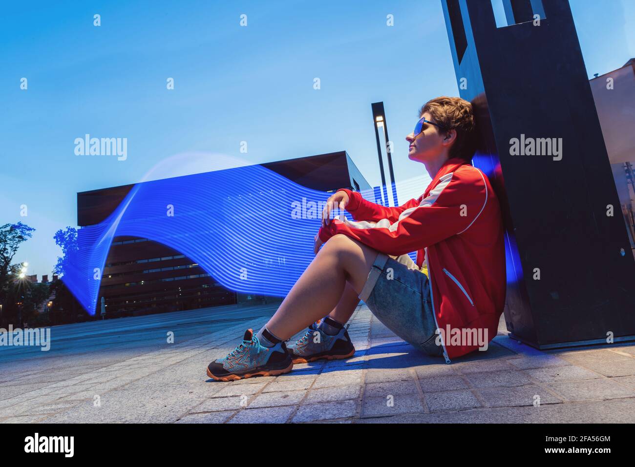 portrait of young woman sitting outdoor on a ground on a long exposure. young woman wearing for night club techno party posing on background with abst Stock Photo