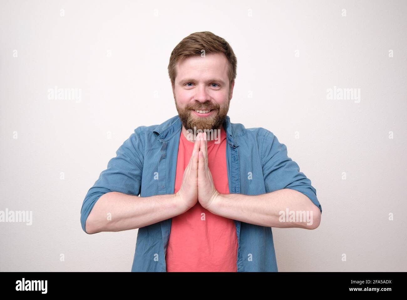 Welcoming, friendly Caucasian man folded his palms together and smiling cutely while looking into the camera. White background. Stock Photo