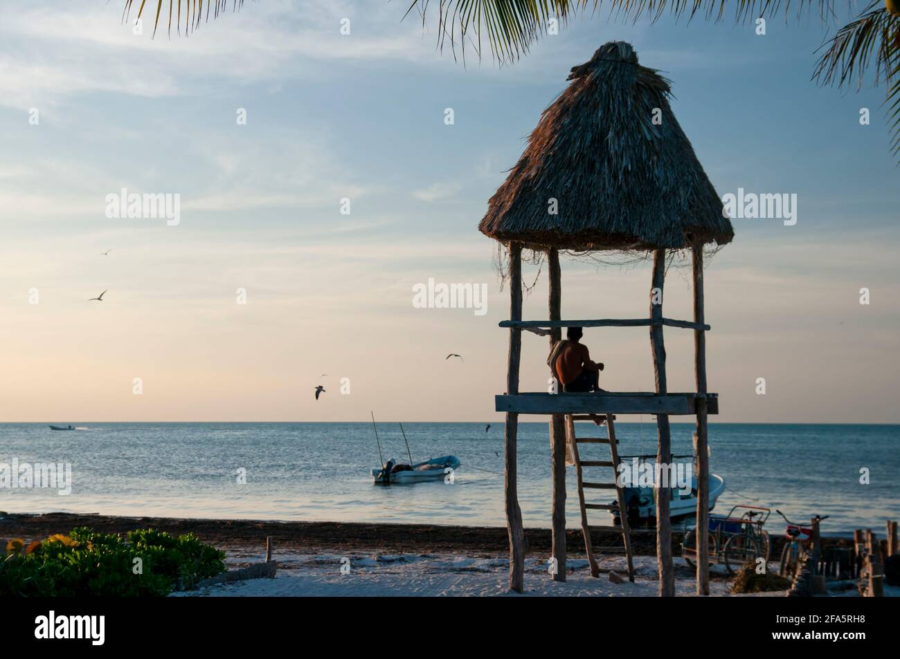 A man sitting in a rustic wooden hut on a beach on the island of Holbox in Mexico observes the horizon over the sea. In the background the Caribbean Stock Photo