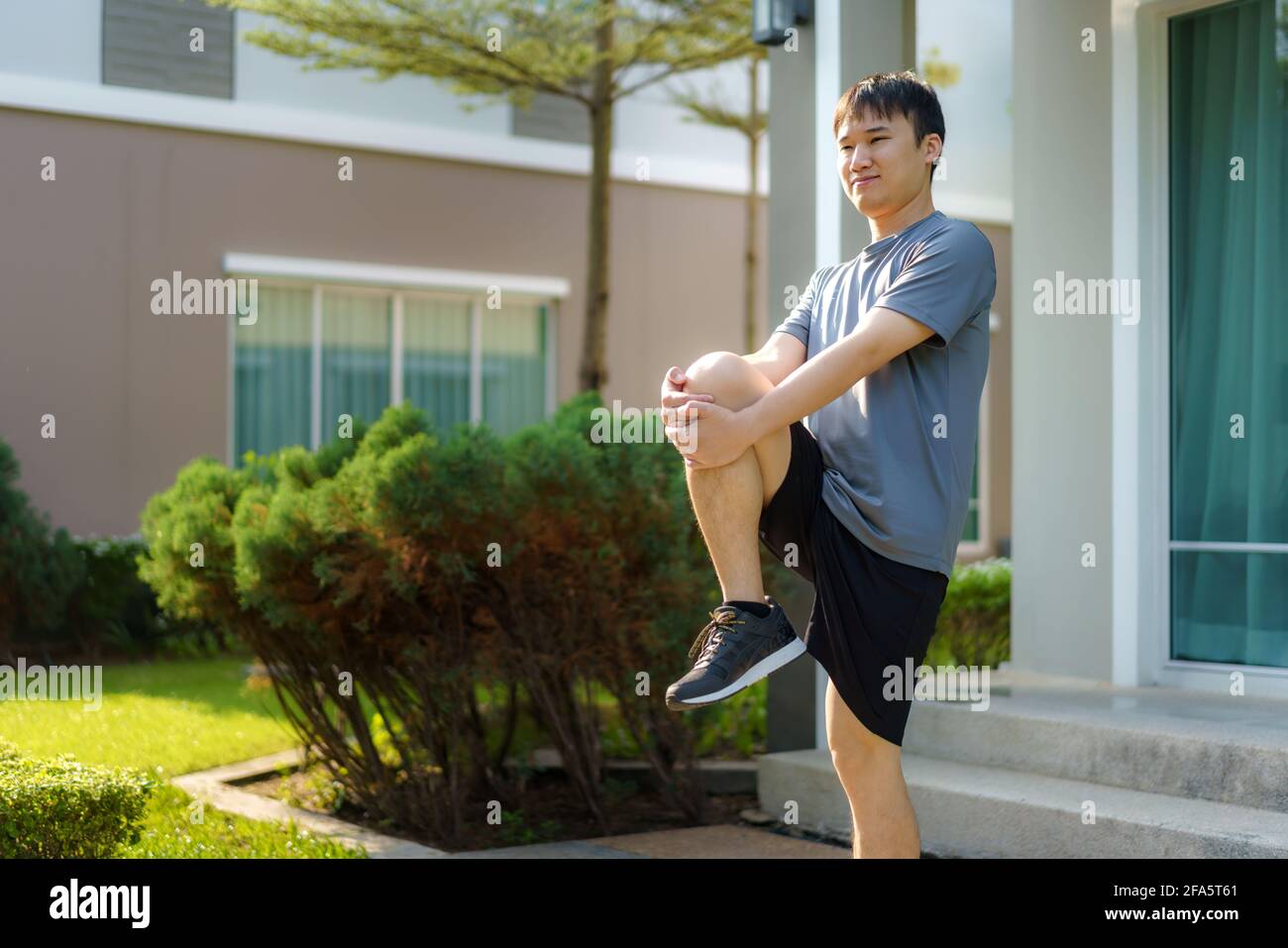 Asian man stretching to warm up or cool down, before or after exercise, near the front door in the neighborhood for daily health and well being, both Stock Photo