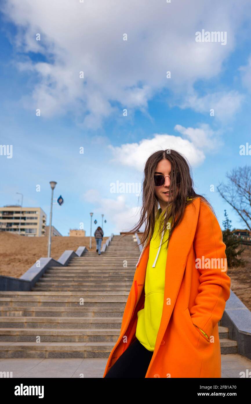 Active good-looking young woman in vivid outfit. Trendy female in  sunglasses, orange coat, yellow hoodie and black jeans against the  background of city stairs and blue sky Stock Photo - Alamy