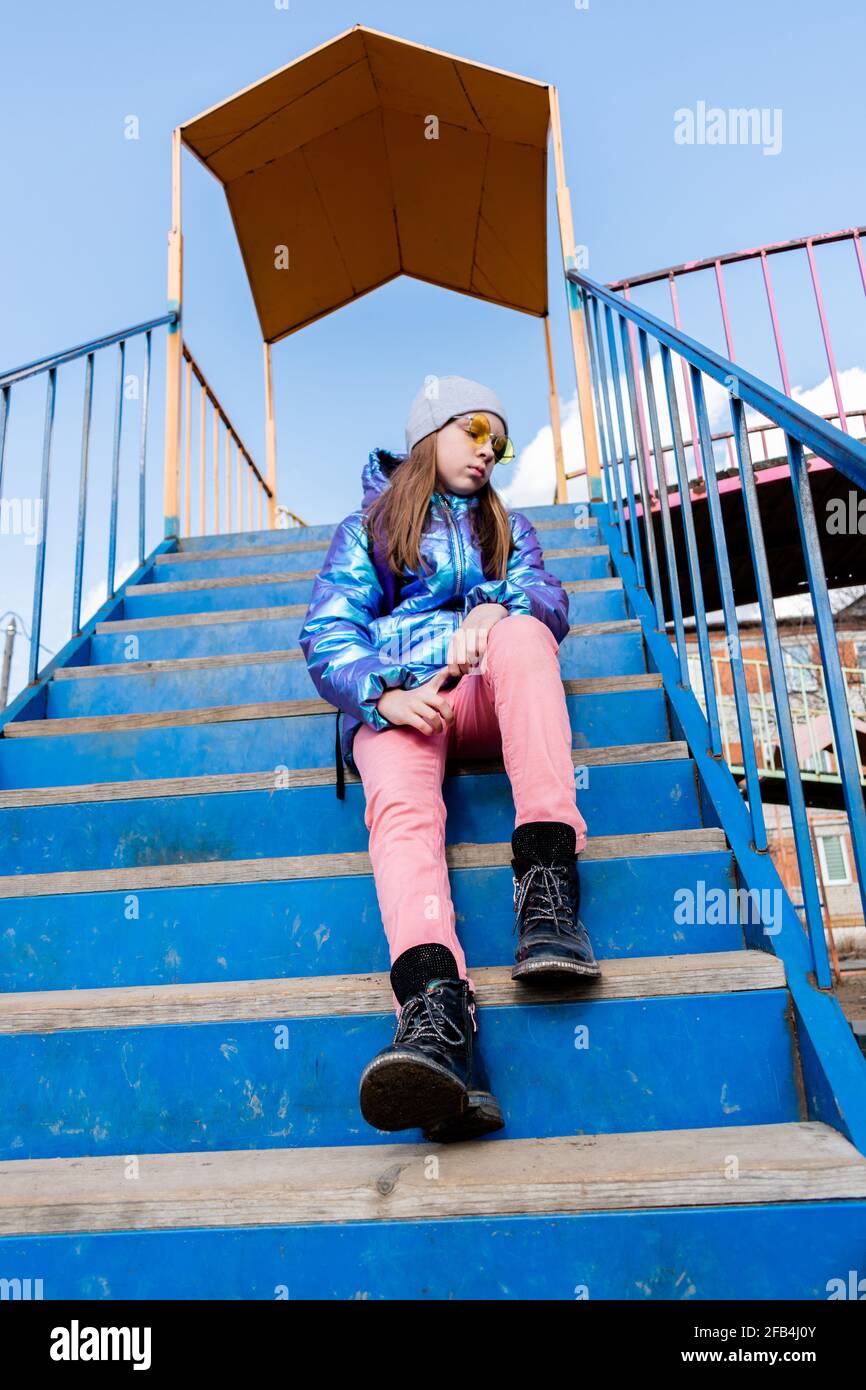 Girl in shiny jacket and sunglasses sits on the stairs. Wellness concept Stock Photo