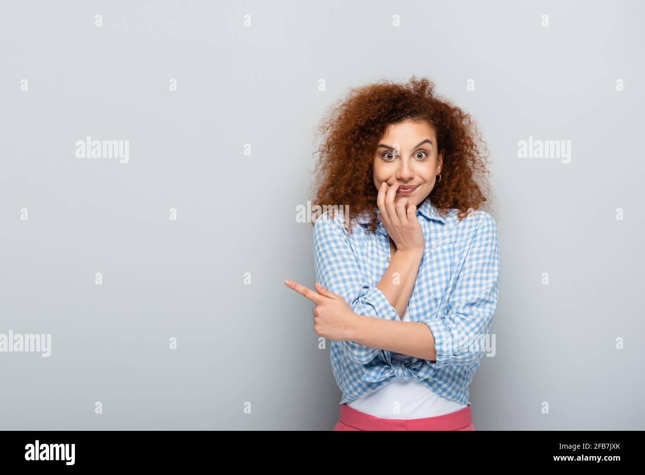 surprised woman pointing with finger while looking at camera on grey background Stock Photo