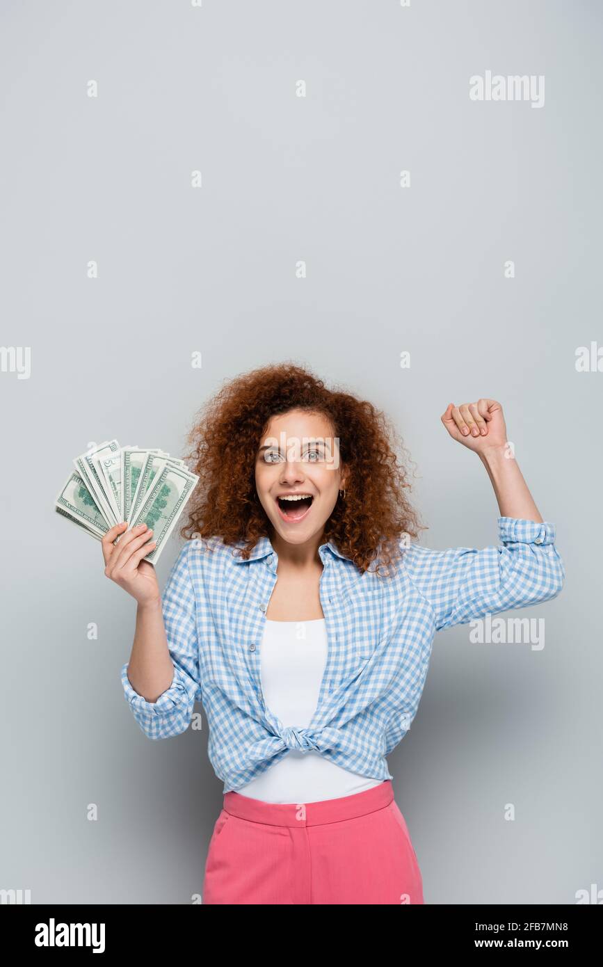 cheerful woman with dollar banknotes showing win gesture on grey ...