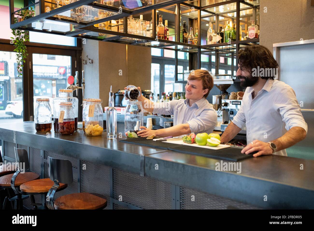 Male trainee putting blackberries in glass jar by bartender at bar counter Stock Photo