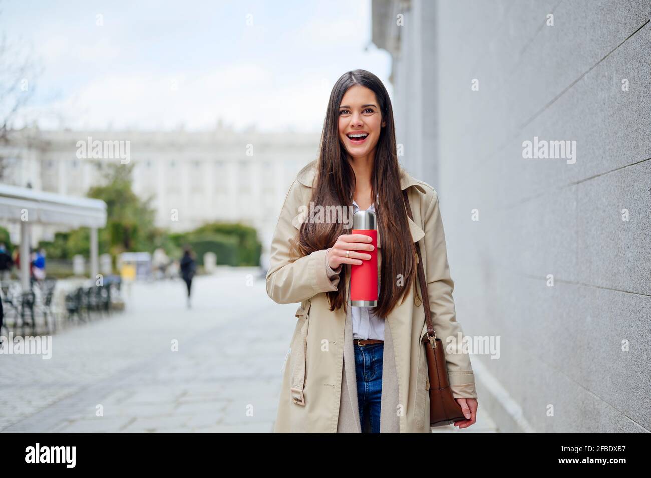 Laughing woman with insulated drink container standing near wall in city Stock Photo