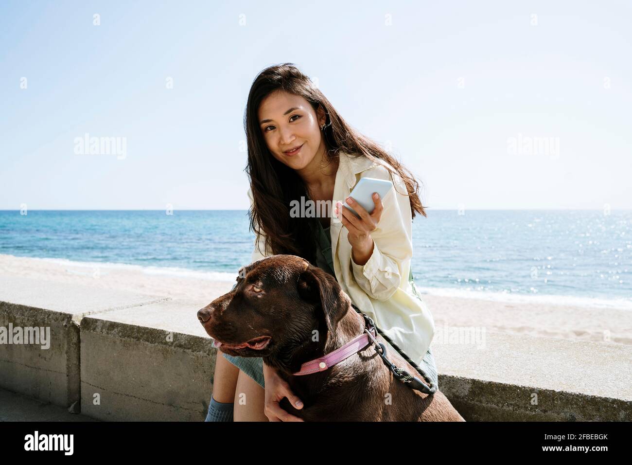 Smiling woman sitting with Labrador Retriever by sea Stock Photo