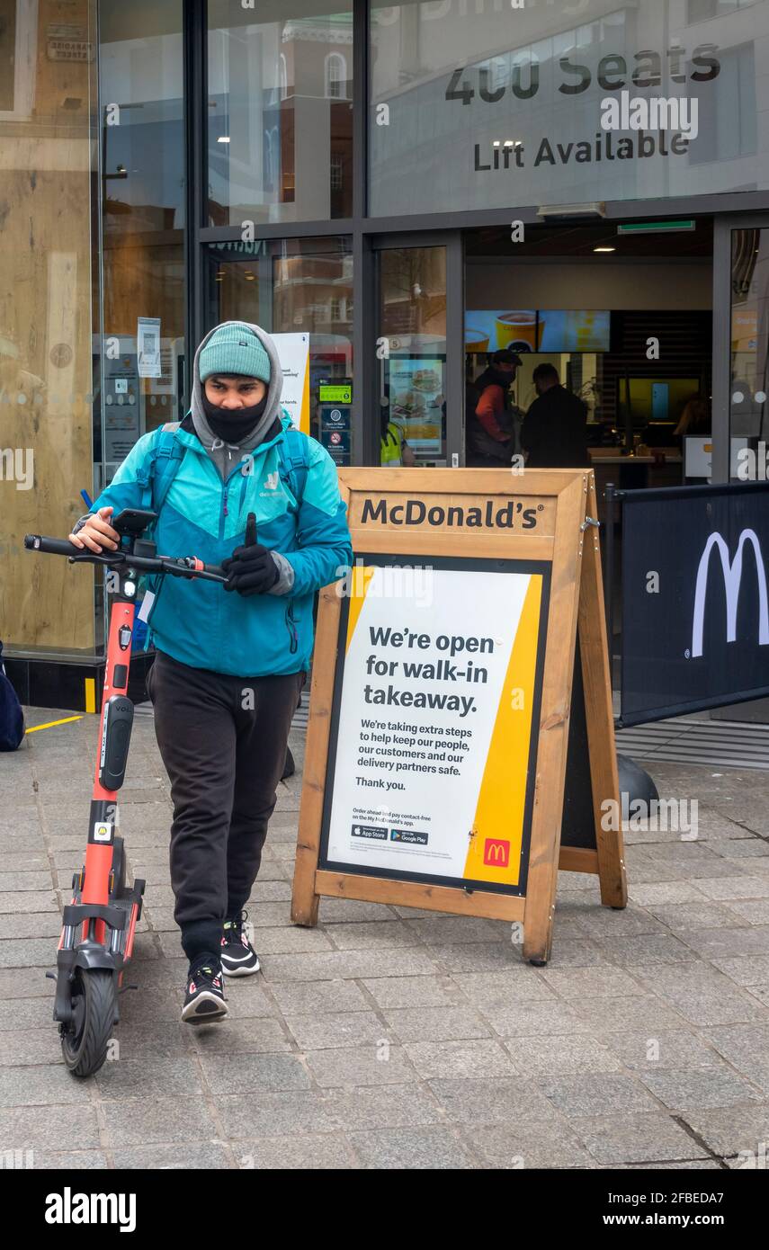 Deliveroo bike delivery courier with face mask leaving McDonald's with a food order. To be delivered on a rented electric scooter. Stock Photo